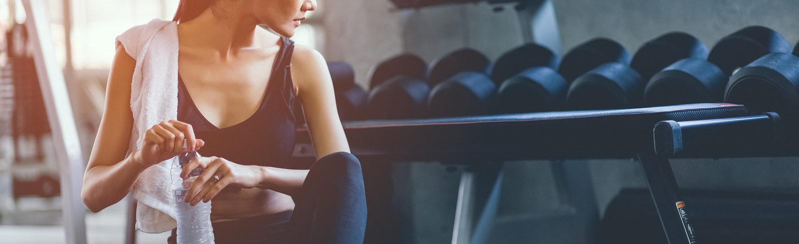 Woman sitting on the gym floor with a water bottle, looking at the weights