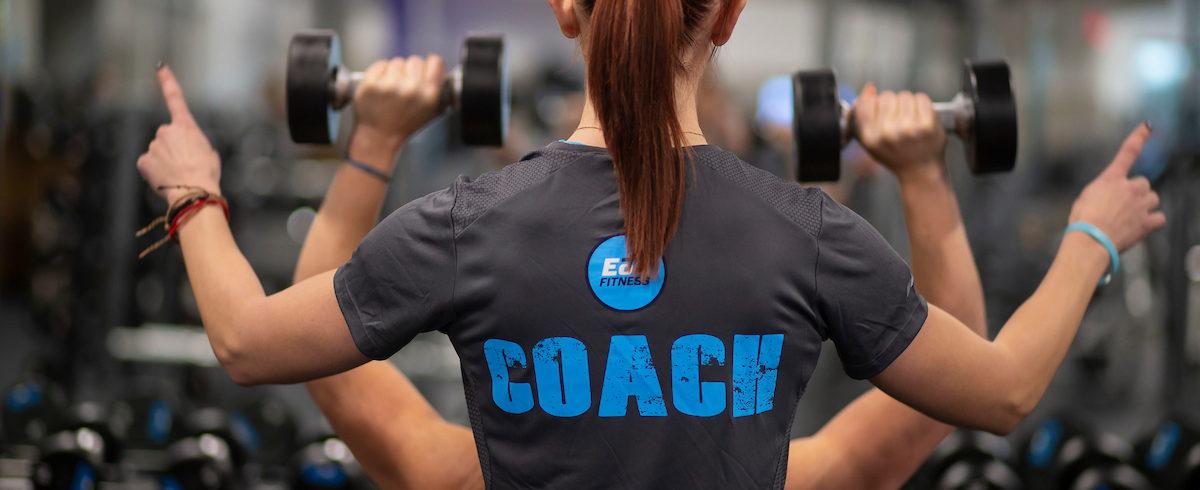 A coach assists a person with dumbbell shoulder presses in a gym