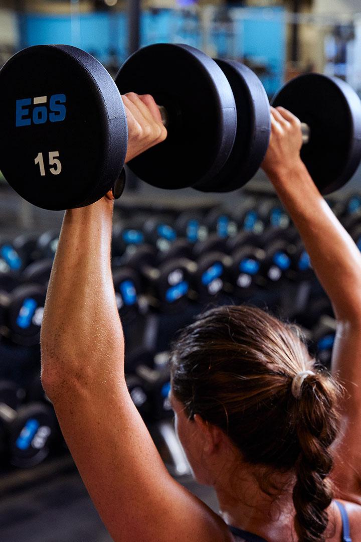 Trainer spotting a woman lifting dumbbells overhead at the gym