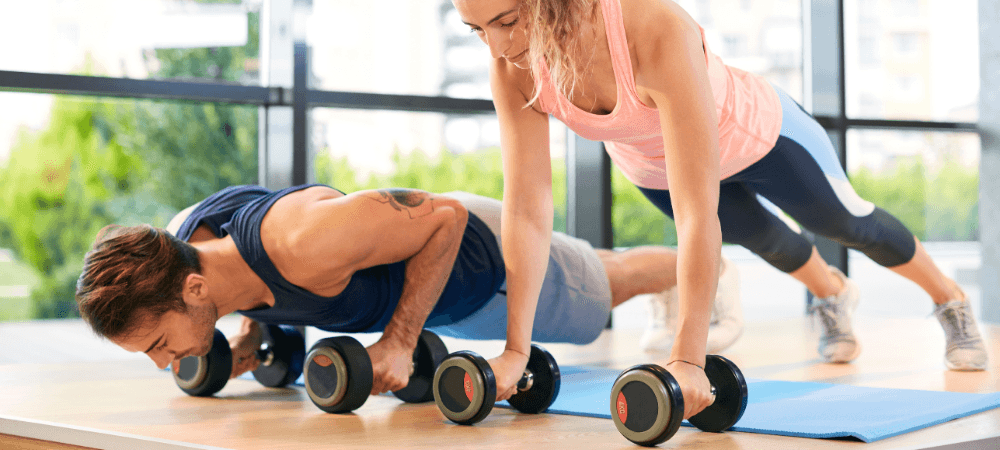 two athletic eos gym members doing dumbbell pushups together in a well lit yoga studio